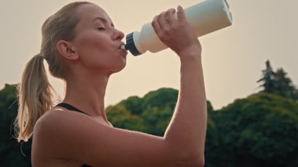 Blondynka sportowiec sportowiec biały biegacz fitness trener czuje się spragniony po porannym treningu jogging napoje czyste filtrowana słodka woda z białej butelki sportowej cieszy się pauzy podczas codziennego treningu - Materiał filmowy, wideo