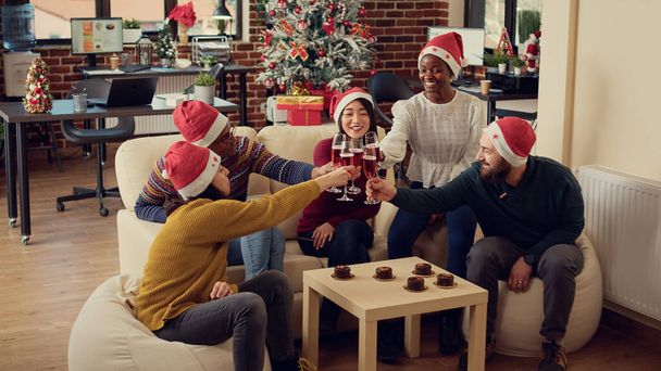Multiethnic group of people clinking glasses of wine during office christmas party, giving toast to celebrate winter holiday. Celebrating seasonal festivity with alcoholic drinks, saying cheers. - 写真・画像