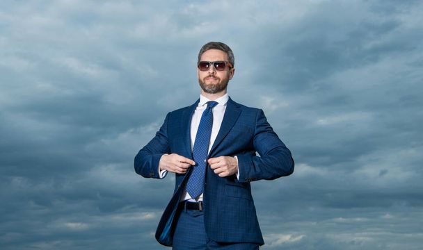 Businessman wearing sunglasses sky background. Grizzled businessman buttoning suit jacket. Serious businessman outdoors. - Фото, зображення