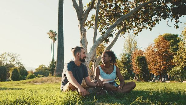 Happy smiling interracial couple talking while sitting on the grass in the park. Backlight - Foto, imagen