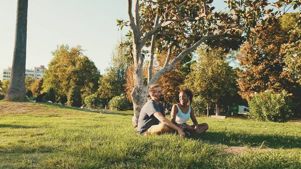 Happy smiling interracial couple talking while sitting on the grass in the park. Backlight - 写真・画像