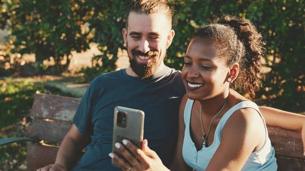 Happy smiling interracial couple taking selfie on phone while sitting on park bench.   - Foto, imagen