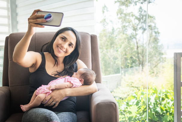 Giovane donna che si fa un selfie seduta su un divano con il suo bambino nel suo salotto - Foto, immagini