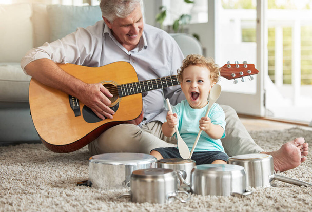 Музыка, кастрюли и детские игрушки со стариком на полу гостиной с кастрюлей и деревянными ложками с гитарой. Память, улыбка и дедушка с бабушкой наслаждаются временем с счастливым внуком. - Фото, изображение