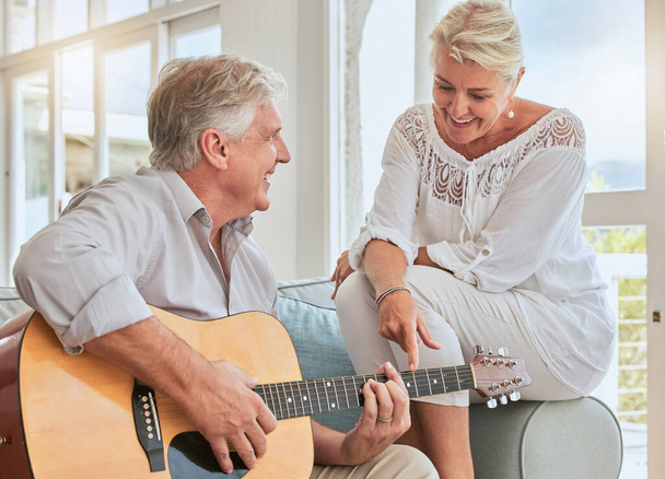 Old couple, guitar and love in home, sofa or couch playing a romantic, lovely or affection musical song for wife. Romance, retired senior man and woman play acoustic string instrument in living room - Photo, Image