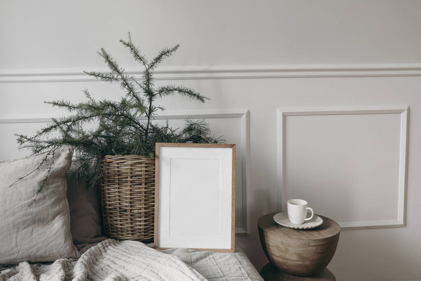 Christmas interior. Blank vertical wooden picture frame mockup on sofa, linen cushions and blanket. Larch, pine tree branches in basket. Cup of coffee on wooden coffee table, white wall background. - Фото, изображение