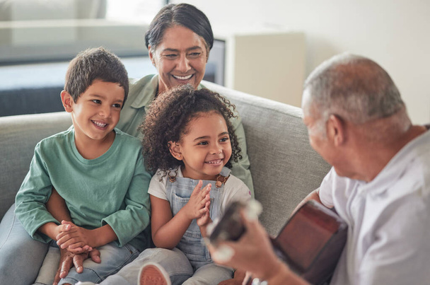 Grandfather playing the guitar for grandmother and children sitting on the sofa in their home. Retired grandparents entertain happy and smiling grandchildren with music on acoustic guitar and bonding. - Photo, Image
