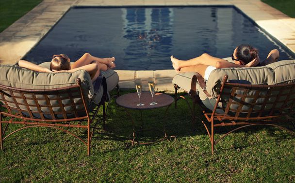 Viviendo la alta vida. Vista trasera de dos mujeres jóvenes relajándose en las tumbonas frente a la piscina - Foto, Imagen