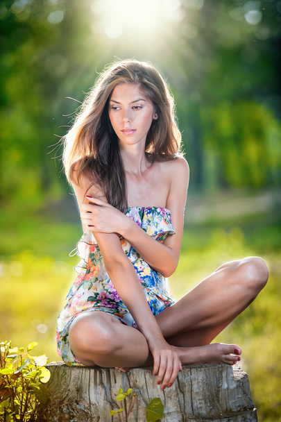 Young beautiful long hair woman wearing a multicolored dress posing on a stump in a green forest. Fashionable sexy attractive girl sitting on hub in a sunny day. Gorgeous girl in garden in summer day. - Φωτογραφία, εικόνα