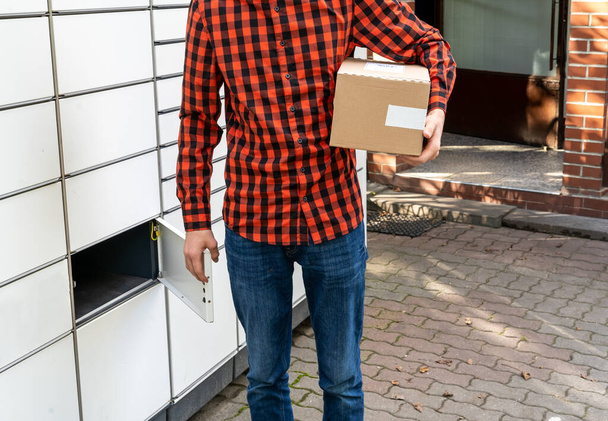 Un homme avec une boîte dans les mains près du terminal de courrier libre-service. Machine de livraison de colis. Personne tenant une boîte en carton. Livraison postale et service postal, achats en ligne, concept de commerce électronique - Photo, image