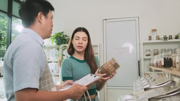 Asian male shopkeeper describes natural organic products to woman customer in refill store, zero-waste and plastic-free grocery, eco environment-friendly, sustainable lifestyles with a reusable shop. - Filmmaterial, Video