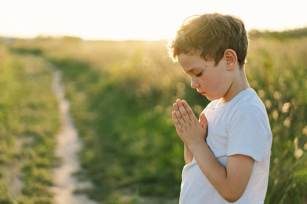 Chłopiec zamknął jej oczy i modlił się na polu o zachodzie słońca. Ręce złożone w koncepcji modlitwy o wiarę, duchowość i religię. - Zdjęcie, obraz