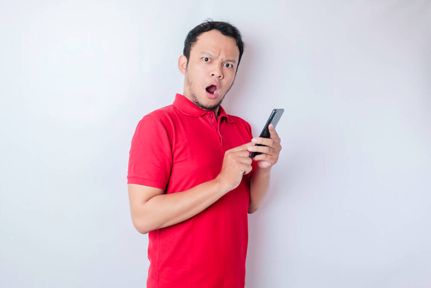 Surprised Asian man wearing red t-shirt pointing at his smartphone, isolated by white background - Foto, Imagen