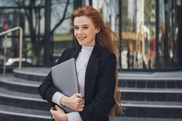 Portrait of a confident businesswoman looking at camera standing with laptop outside near office building. Caucasian female business person. Stylish - Photo, Image