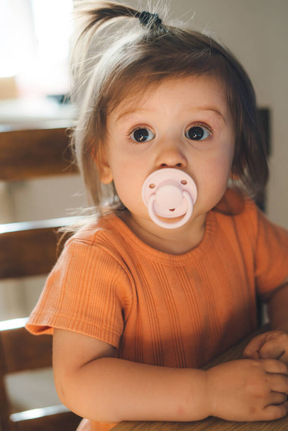 Portrait of adorable baby girl with big brawn eyes sucking a pacifier, sitting on wooden chair at kitchen. Happy little girl at home. Explore world. - Foto, imagen