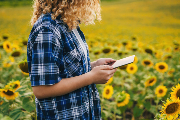 Gekropte weergave van boer vrouwelijke handen houden tablet staande in zonnebloemenveld op hete zomermiddag. Buiten levensstijl. Gecultiveerd landbouwgebied - Foto, afbeelding