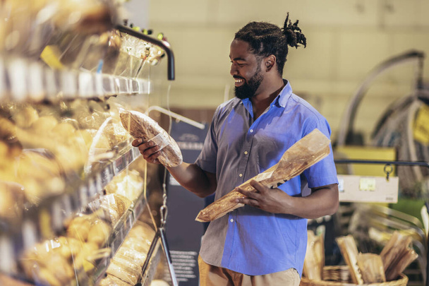 Hombre guapo está eligiendo pasteles y sonriendo mientras hace compras en el supermercado - Foto, imagen