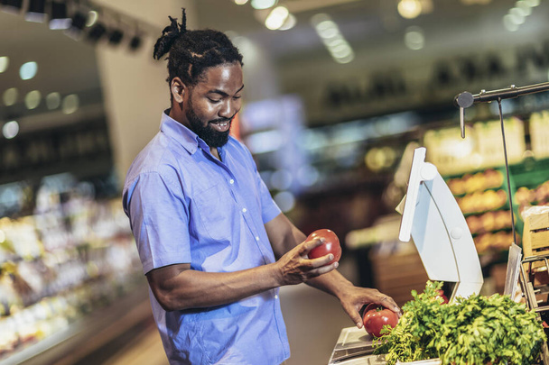 Αφρο-Αμερικανός άνθρωπος μετρά φρούτα και λαχανικά σε ψηφιακή κλίμακα για τους πελάτες. - Φωτογραφία, εικόνα