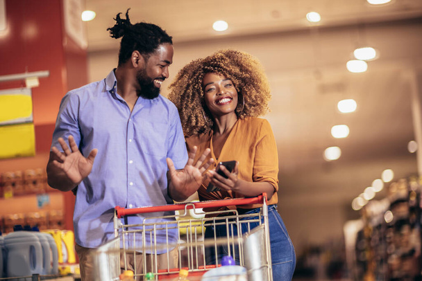 African American couple choosing products using phone during grocery shopping in modern supermarket - Foto, afbeelding