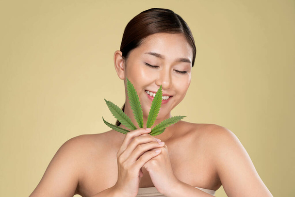 Portrait of young ardent girl with healthy fresh skin holding green hemp leaf. Combination of beauty and cannabis concept. - Фото, изображение