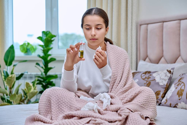 Young teenage female sitting in bed with runny nose spray drops, sneezing into tissues, burying her nose. Autumn winter, rhinitis runny nose stuffy nose, treatment, flu season - Foto, immagini