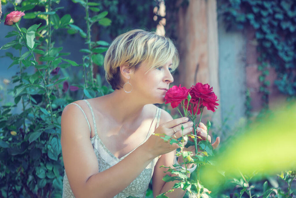 Beautiful pensive woman smelling red roses and enjoying in her garden. - Valokuva, kuva