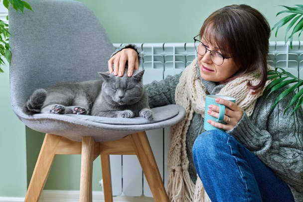 Cold winter season woman with cat near heating radiator. Middle-aged female in warm woolen knitted sweater with cup of hot drink and pet lying on chair. Heating season, lifestyle, crisis energy saving - Foto, Bild