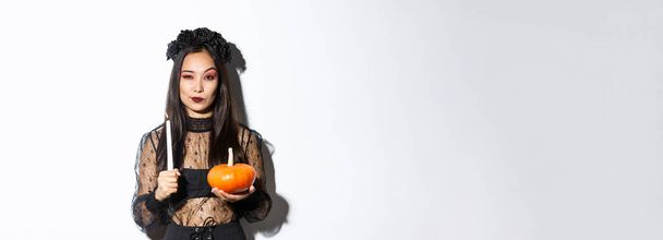 Image of beautiful asian woman in witch costume, holding lit candle and pumpkin, celebrating halloween. - Foto, Imagem