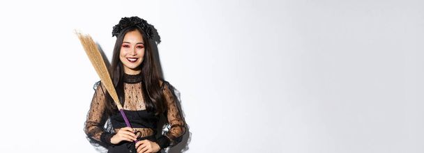 Portret van een glimlachende mooie Aziatische vrouw in heksenkostuum die bezem vasthoudt en blij kijkt naar de camera, Halloween viert, geniet van truc of traktatie, witte achtergrond. - Foto, afbeelding