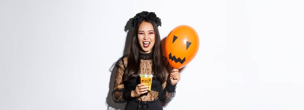 Τρομακτικό μαγικό κόλπο ή κέρασμα στις Απόκριες, κρατώντας γλυκά και πορτοκαλί μπαλόνι, στέκεται σε γοτθικό φόρεμα δαντέλα με μαύρο στεφάνι. - Φωτογραφία, εικόνα