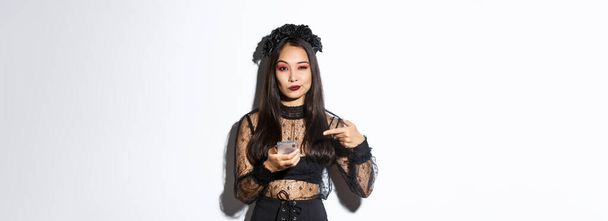 descarada hermosa mujer asiática en vestido gótico y corona negra señalando el dedo en el teléfono móvil, mostrando algo acerca de halloween. - Foto, imagen
