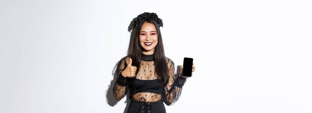 Immagine di donna asiatica felice e soddisfatta in costume di Halloween mostrando pollici in su e dimostrando schermo del telefono cellulare, sorridente soddisfatto, in piedi su sfondo bianco. - Foto, immagini