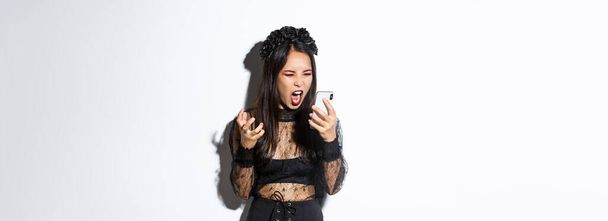 Porträt einer wütenden asiatischen Frau im Halloween-Kostüm, die verrückt aussieht, ihr Handy anbrüllt und wütend grimmig vor weißem Hintergrund steht. - Foto, Bild