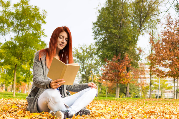 Young redhead woman reading book and relaxing in autumn day at the park.  - 写真・画像