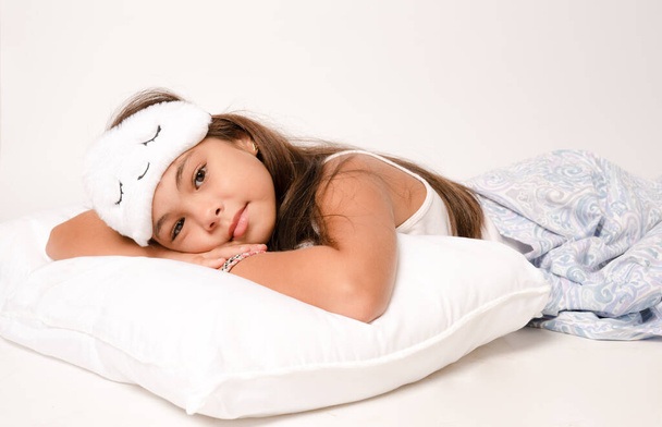 Portrait of cute girl wearing sleeping mask lying in the bed isolated over white background. - Photo, Image