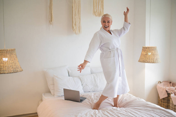 Full-size portrait of a merry mature woman in the terry bathrobe dancing on the bed - Foto, Imagem