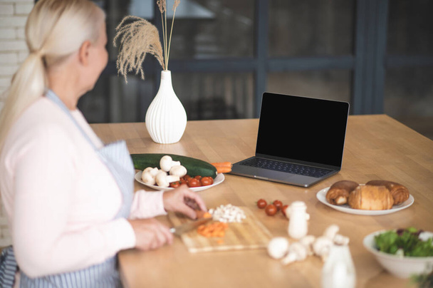 Blonde Frau im Kittelschurz schneidet die Möhren auf dem Holzbrett, während sie auf den Laptop-Bildschirm schaut - Foto, Bild