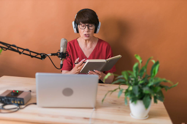 Portrait of mature woman wearing headphones and talking at online radio station - podcast and broadcast - 写真・画像