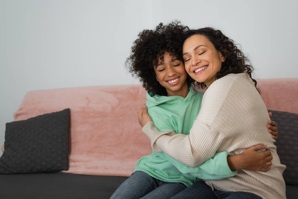 Χαρούμενη Αφροαμερικανή μητέρα και κόρη αγκαλιάζονται ενώ κάθονται στον καναπέ. - Φωτογραφία, εικόνα