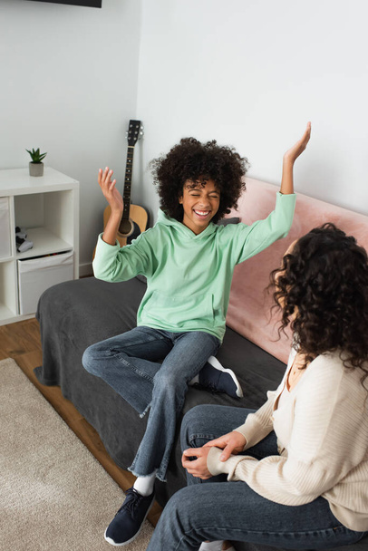 happy african american girl smiling while gesturing near curly mother in living room  - Zdjęcie, obraz