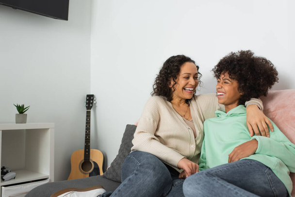 curly african american mother hugging cheerful preteen daughter in living room  - Photo, Image