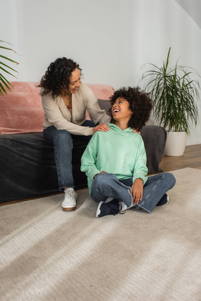 curly african american mother looking at excited preteen daughter sitting with crossed legs on carpet  - Foto, immagini
