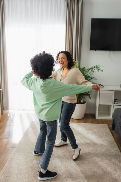 animado afro-americano mulher de mãos dadas com encaracolado preteen filha enquanto dança em casa - Foto, Imagem