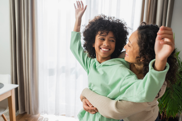 cheerful african american woman hugging happy preteen daughter with raised hands  - Photo, image