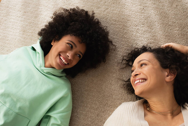 vue du haut de heureuse fille afro-américaine souriant tout en regardant la mère gaie et couché sur le tapis  - Photo, image