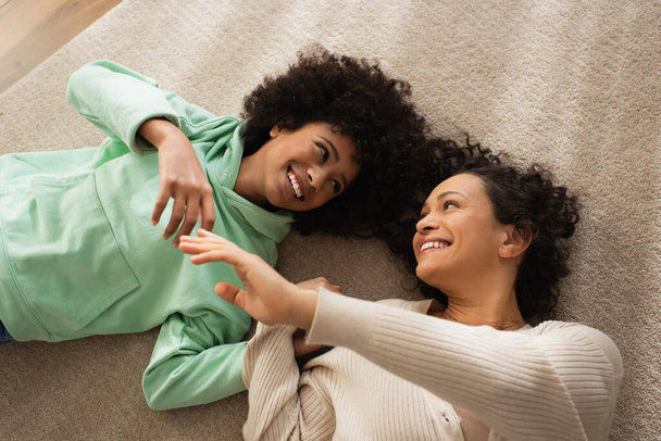 top view of joyful african american girl smiling while lying on carpet with cheerful mother  - 写真・画像