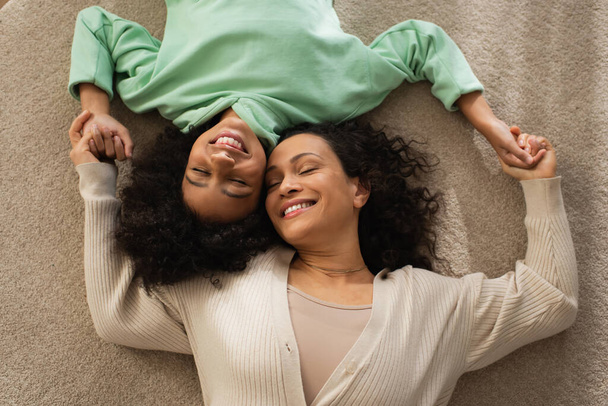 top view of smiling african american girl lying on carpet with cheerful mother  - Фото, зображення
