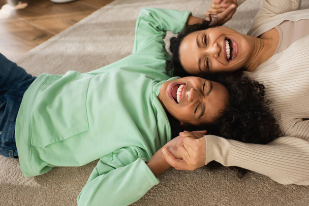 positive african american girl laughing while lying on carpet with cheerful mother  - Foto, Imagem