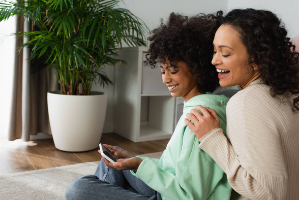 happy african american preteen girl sitting on carpet and holding smartphone near excited mother - Foto, imagen