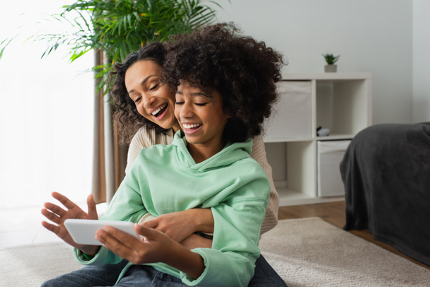 happy african american preteen girl and curly mother using mobile phone in living room - Zdjęcie, obraz